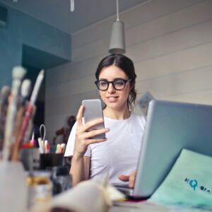 A woman using her phone at a desk, surrounded by art supplies and a laptop, in a creative workspace.