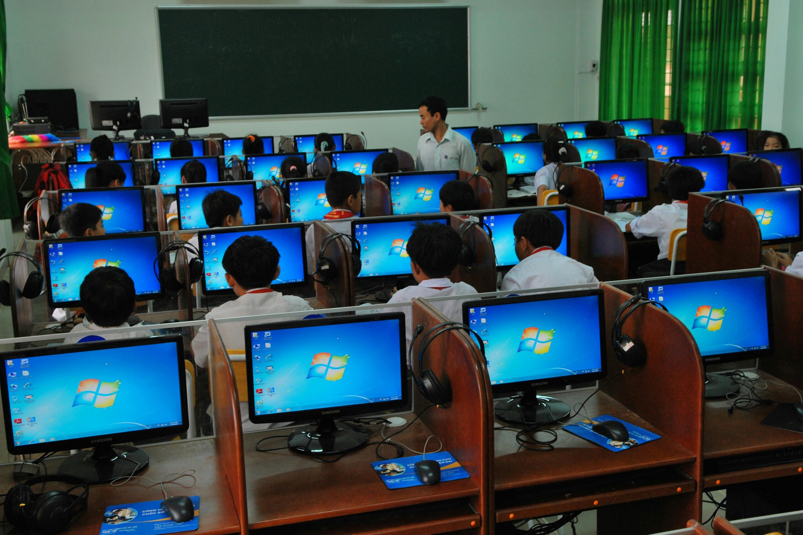 Home Students learning in a computer lab with multiple monitors and a teacher instructing.