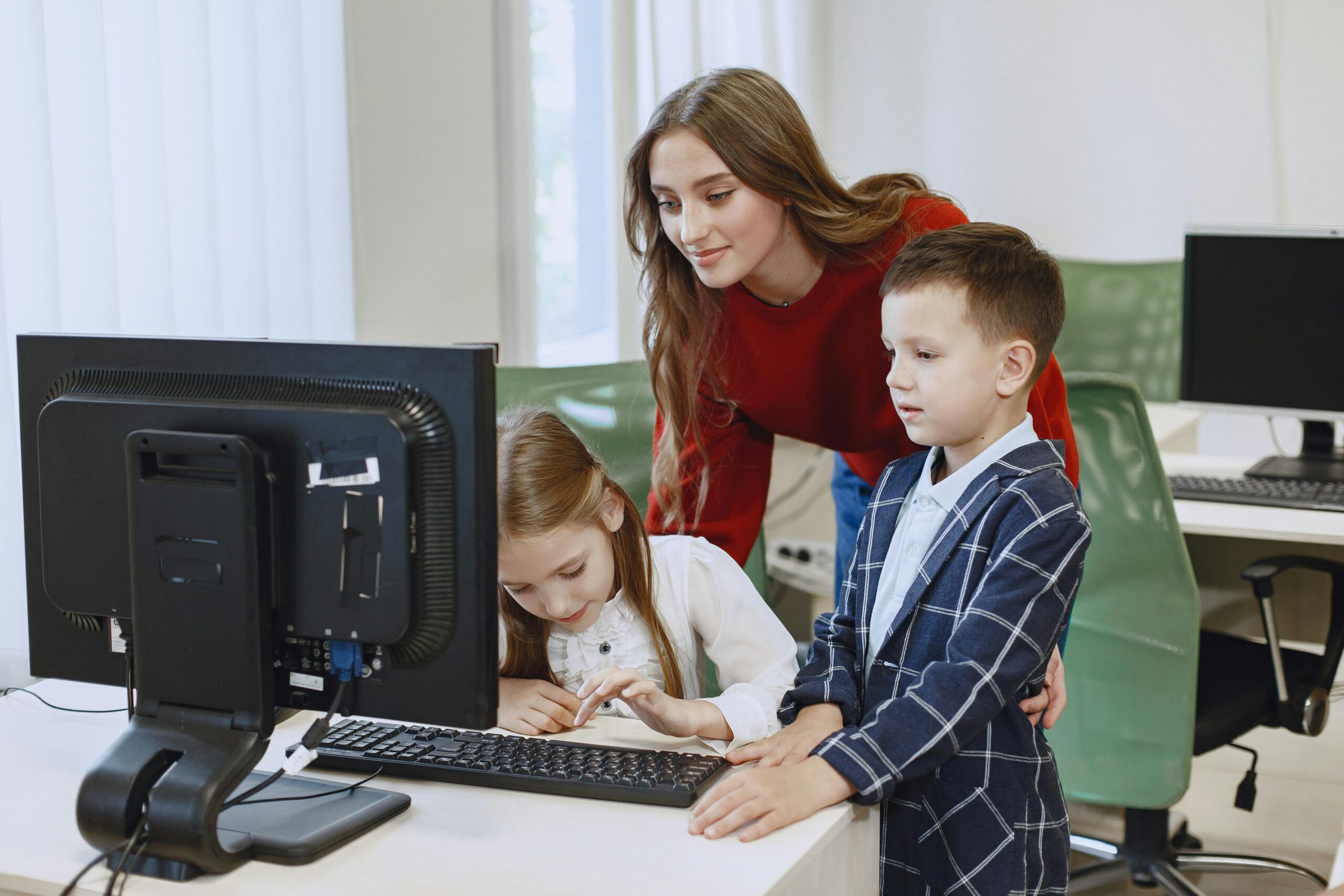 Home A teacher guides two children using a computer in a classroom setting.